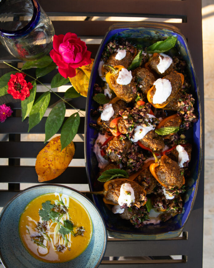 A colorful and vibrant dish featuring roasted baby sweet peppers stuffed with a flavorful Moroccan-spiced ground lamb mixture, garnished with dollops of creamy coriander yogurt, olive tapenade, and fresh mint leaves.