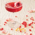 A red pomegranate cosmo as a Pretty Woman inspired drink sits in a tall martini glass with an edible rose floating on top and pomegranate seeds and an orange twist laying on the white table below.