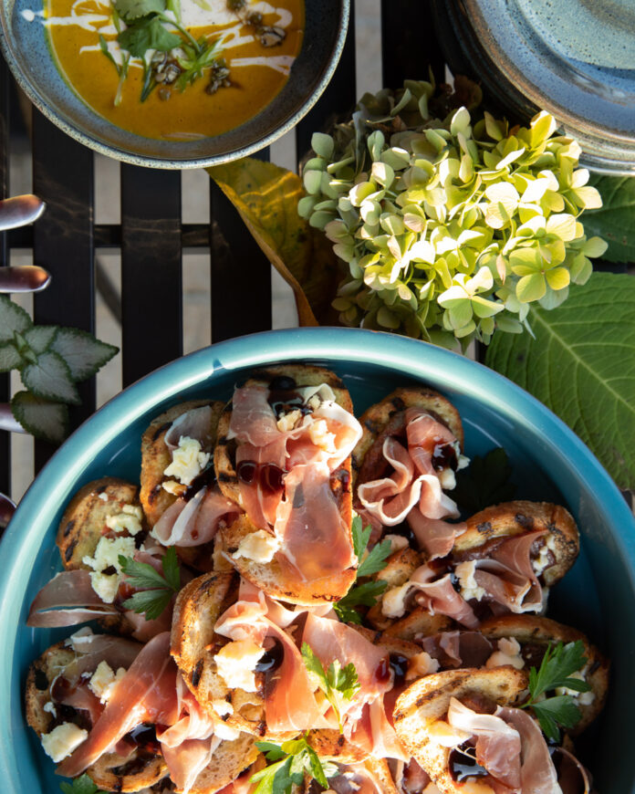 A plate featuring slices of toasted sourdough bread topped with thin slices of prosciutto, dollops of apple butter, shredded aged cheddar cheese, and drizzles of balsamic glaze and olive oil.