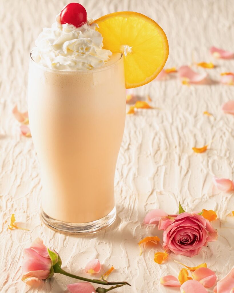 An orange creamsicle shake with whipped cream, a cherry on top, and an orange wheel garnish from Say Anything on a white table with rose petals.