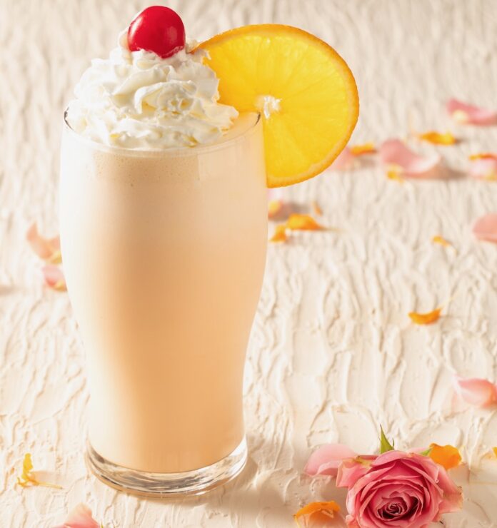 Say Anything Orange Creamsicle Shake An orange creamsicle shake with whipped cream, a cherry on top, and an orange wheel garnish from Say Anything on a white table with rose petals.