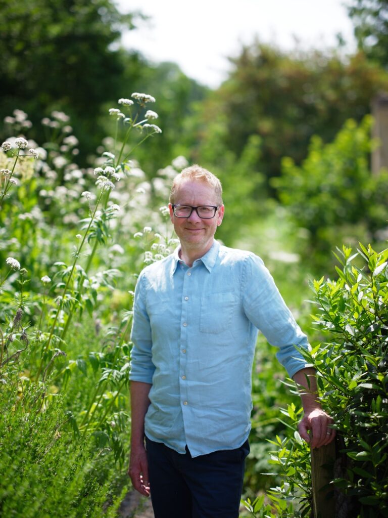 Mark stands in a green field, smiling in a blue button down shirt and black pants.
