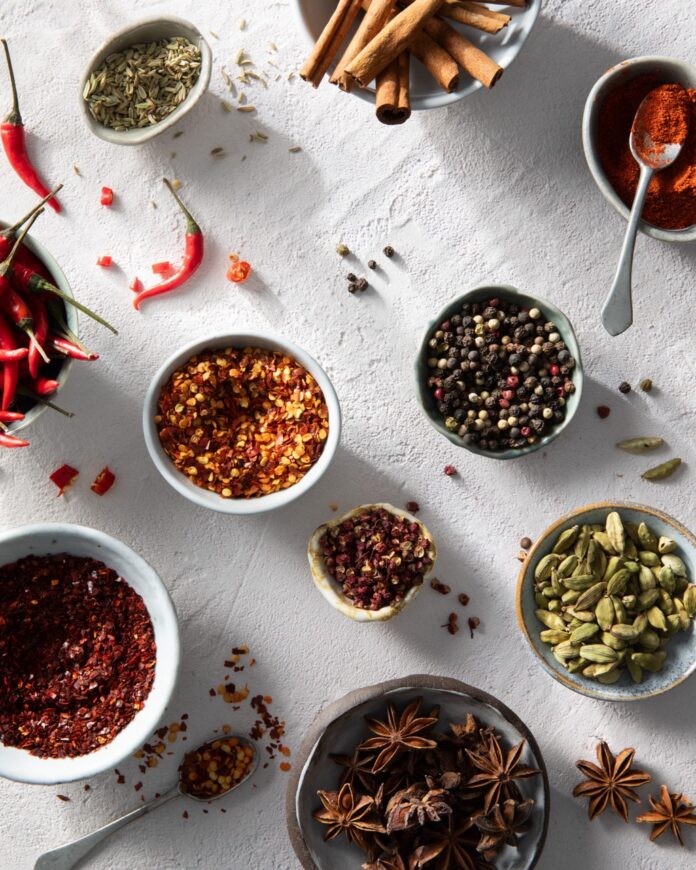 Various small white bowls of red and green spices sit amongst a white table with chili peppers and seeds scattered about.