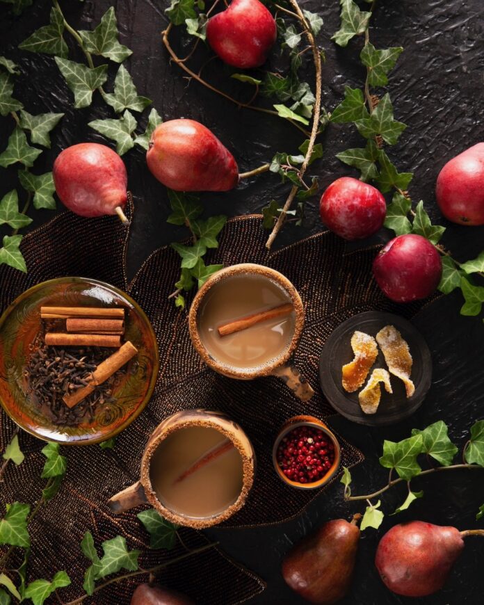 Two brown megs with coffee and a cinnamon stick inside sit next to a plate of cinnamon sticks and surrounded by red fruits on a green vine.