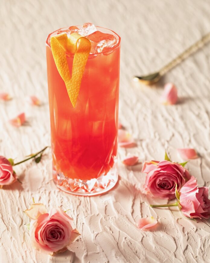 Sweet Home Alabama Slammer In a tall glass sits a red Sweet Home Alabama Slammer garnished with a heart formed orange peel and surrounded by pink roses on the table.