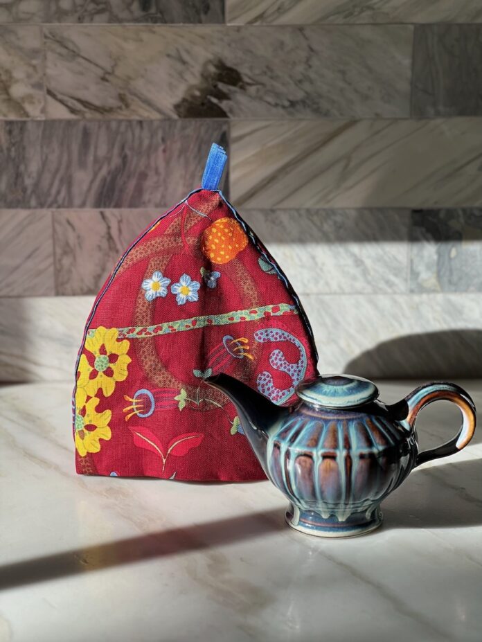 A red embroidered tea cozy staged with a cup of tea on a marble countertop