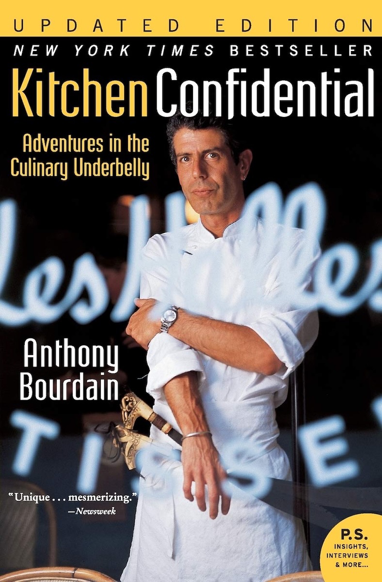 The cover of Anthony Bourdain's Kitchen Confidential, with Bourdain standing in front of a black background wearing a chef's outfit. 