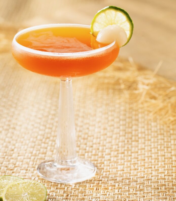 A tall martini glass holds an orangish thai getaway drink in honor of The White Lotus season with a lime wheel garnish.