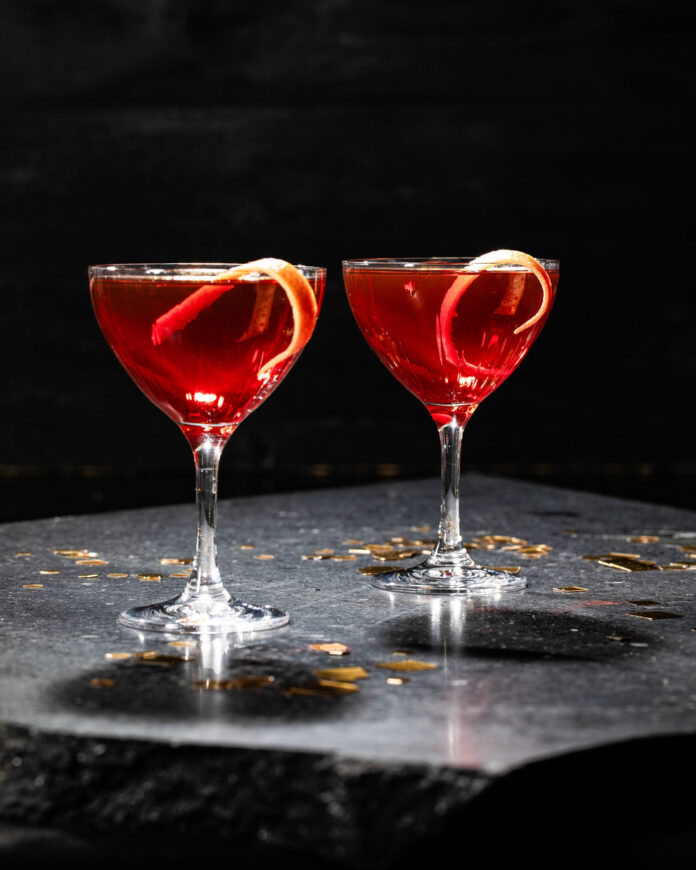 The Palazzo Two elegant glasses of red wine garnished with a twist of grapefruit, set against a cozy indoor backdrop.