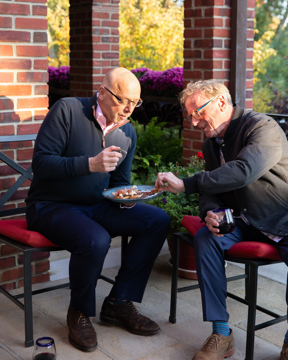 Two older gentlemen enjoying a meal together on the patio of a home, surrounded by lush greenery and brick architecture.