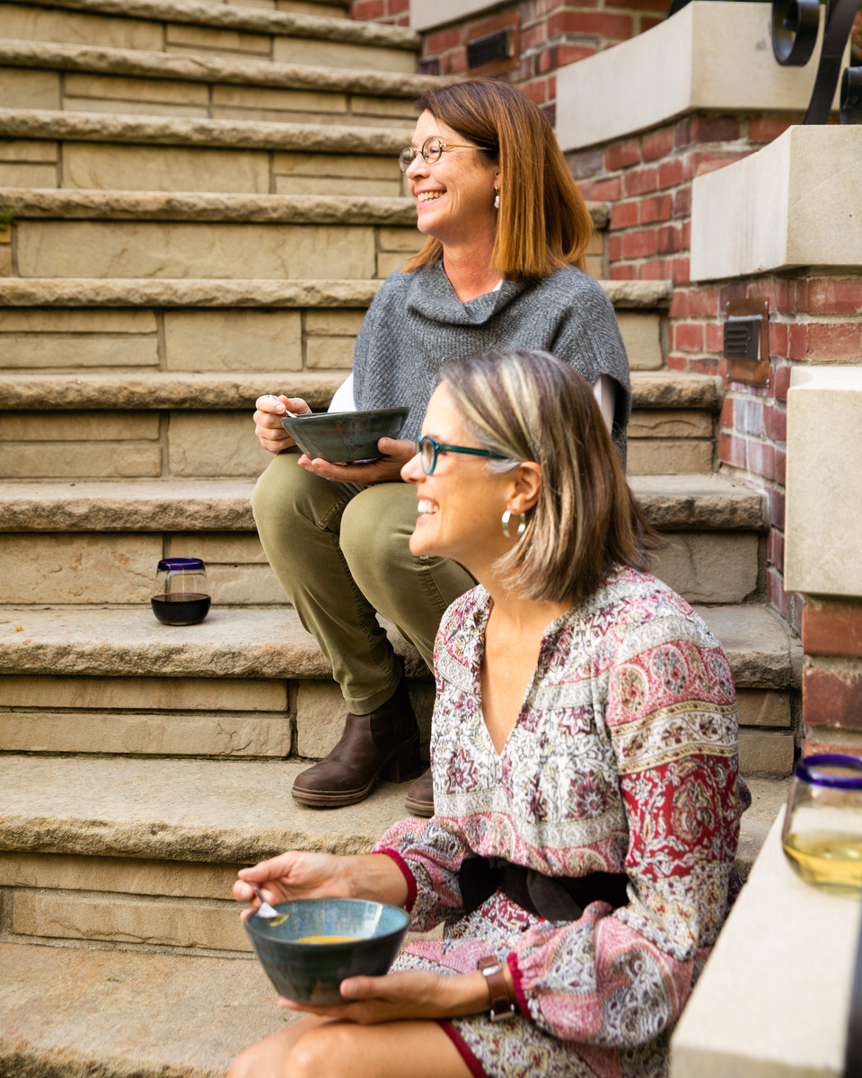 Two women sitting on stone steps, enjoying a meal together while conversing and laughing.