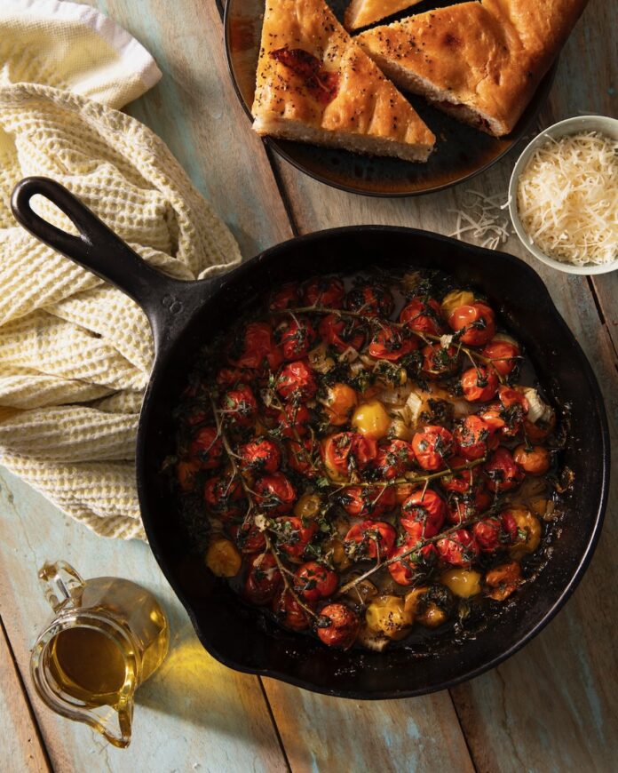 In a cast iron skiller sits a hot cherry tomato salad with cherry tomatoes and herbs as focaccia bread, cheese, and oil sit off to the side.