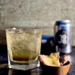 A short rocks glass holds a Horsefeather cocktail for the Kansas City Chiefs with a small bowl of ginger slices in front of it and cans of ginger beer behind it.