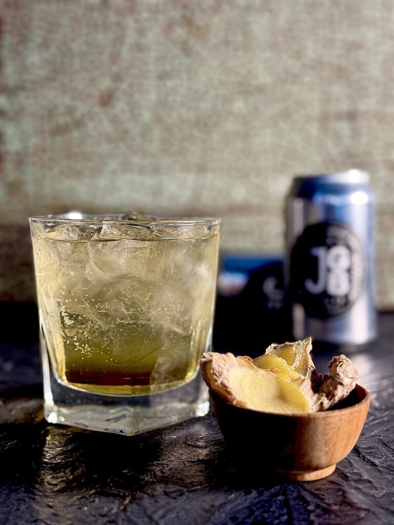 A short rocks glass holds a Horsefeather cocktail for the Kansas City Chiefs with a small bowl of ginger slices in front of it and cans of ginger beer behind it.