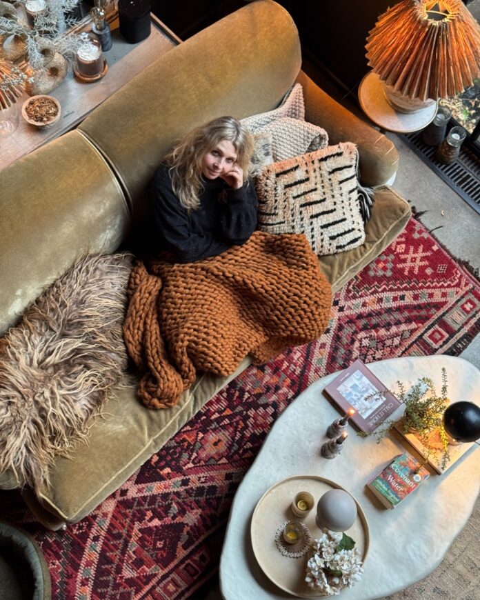 Abigail Ahern sits on a brown couch surrounded by maximalist design elements like various patterned pillows, a bold red accent rug, lamps, and other accessories.
