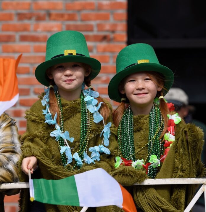 Two little girls in St. Patrick's Day green hats, beads, and green sweaters while one holds an Irish flag.