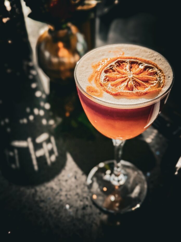 A Nickel Boys inspired cocktail sits on a dark table with a ray of light shining through as a lemon slice sits on top of the cocktail.
