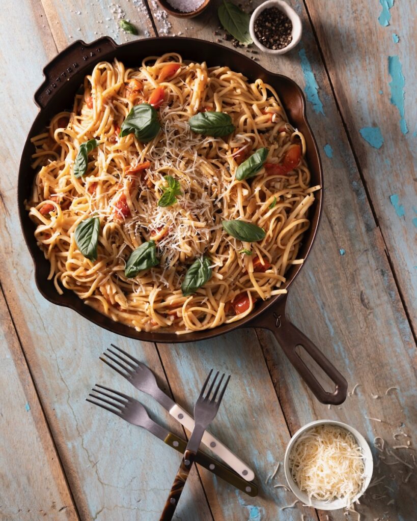 In a skillet pan sits a large servings of pasta with tomatoes, basil, and cheese on top. It all sits on a picnic table with forks and a small container of parmesan underneath the pan.
