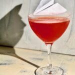 A Paper Plane Cocktail with an actual paper plane garnish sits on a white and blue background.