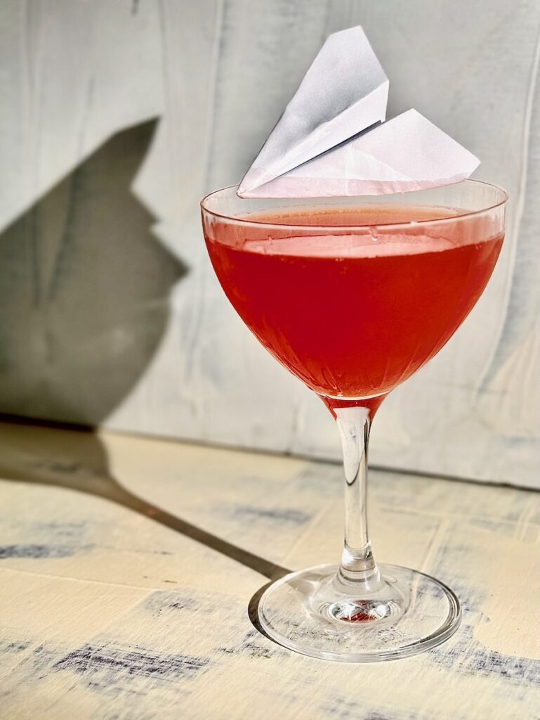 A Paper Plane Cocktail with an actual paper plane garnish sits on a white and blue background.