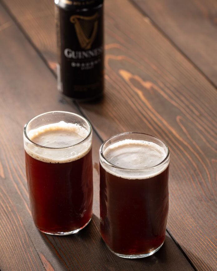 Two short glasses hold Snakebite cocktails which sit on a wooden table with a can of Guinness sitting behind them.