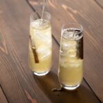 Two glasses of pale yellow tepache with a cinnamon stick in them on a brown background.