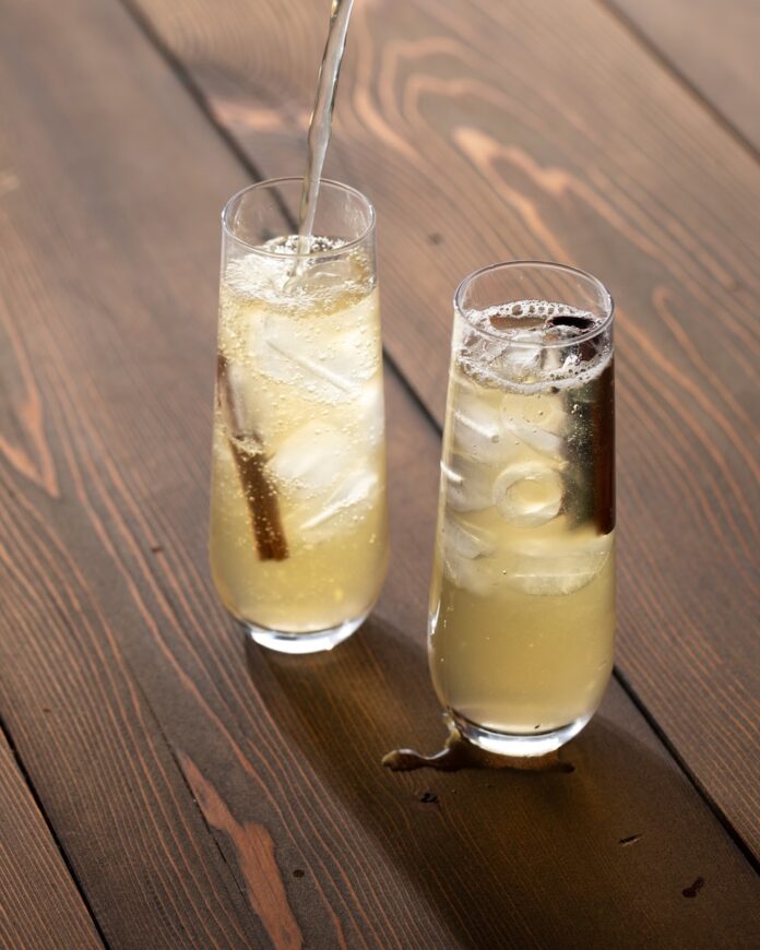 Two glasses of pale yellow tepache with a cinnamon stick in them on a brown background.