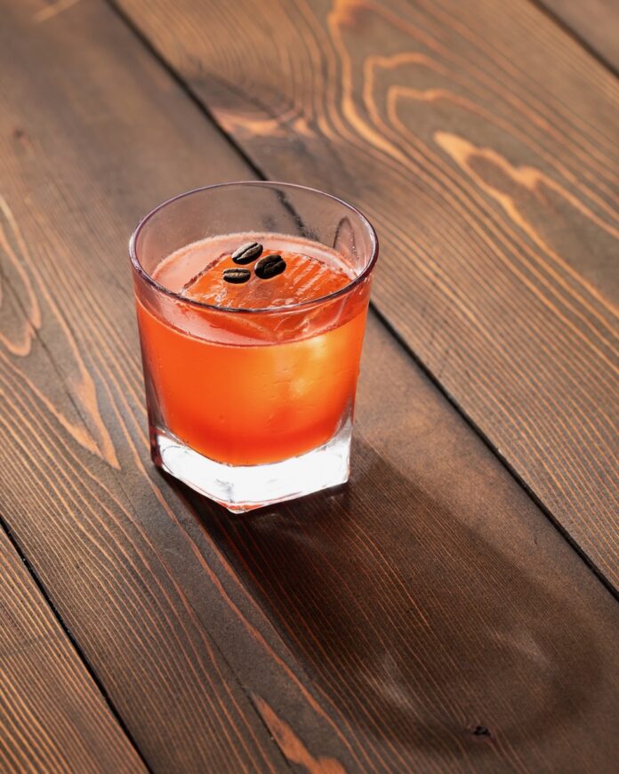 A short cocktail glass with an orange Conclave cocktail with one large ice cube inside and coffee beans on top of it, with the glass sitting on a wood picnic table.