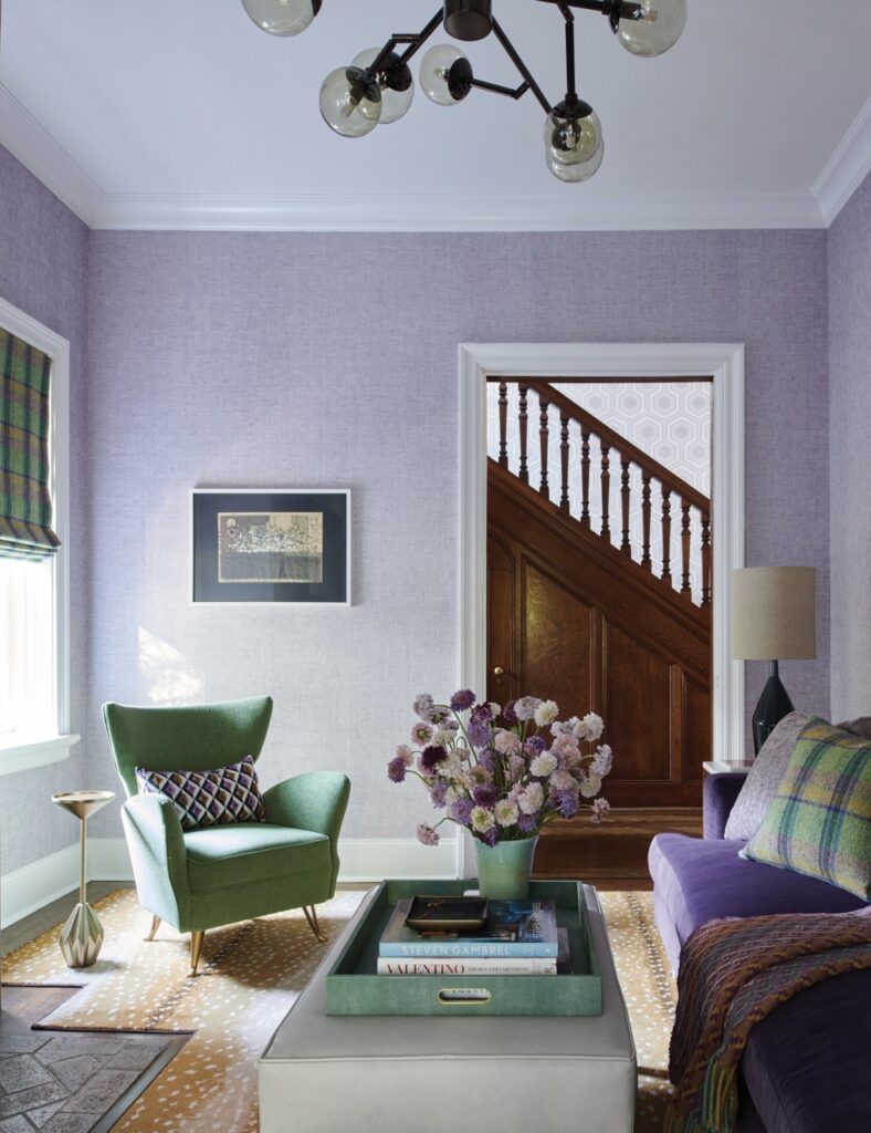 A lavender wall closes in a space with a green reading chair and couch with a pot of flowers on the coffee table.