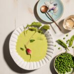 A white bowl holds a Chilled Pea Soup with radish and mint garnishes as a bowl of peas sits off to the right.