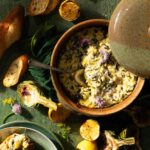 A brown bowl full of Dandelion Artichoke Dip with a spoon in it and crusty bread laying around the dish.