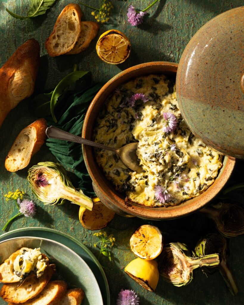 A brown bowl full of Dandelion Artichoke Dip with a spoon in it and crusty bread laying around the dish.