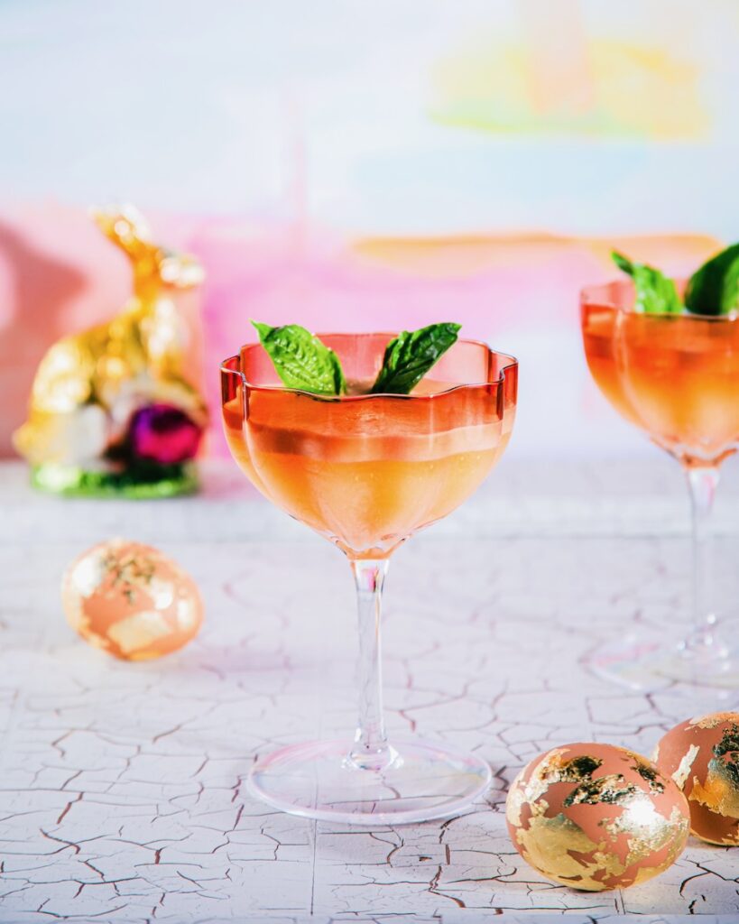 A coupe glass holds a carrot cocktail with two basil leaves as bunny ear garnishes and golden eggs around the glasses.