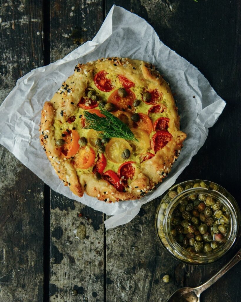 An Everything Bagel Tomato Tartelette sits on a piece of parchment paper with a jar of capers sitting nearby.