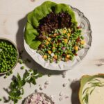 A white bowl holds butter lettuce leaves with plenty of peas, cheddar cheese, and bacon. Off to the bottom right corner sits a bowl of green goddess dressing.