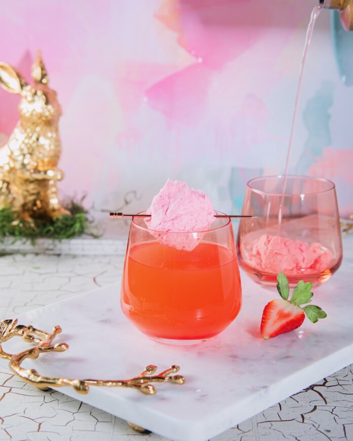 A short wine glass with a Here Comes Peter Cottontail inside as a ball of cotton candy sits over the top of the glass. A golden bunny sits in the background by a pastel backdrop.