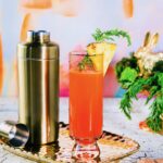 A tall glass holds a Hoppin’ Down the Bunny Trail with a Buzzed Bunny Mimosa in an orange color with a pineapple slice garnish as the whole glass sits beside a gold cocktail shaker.