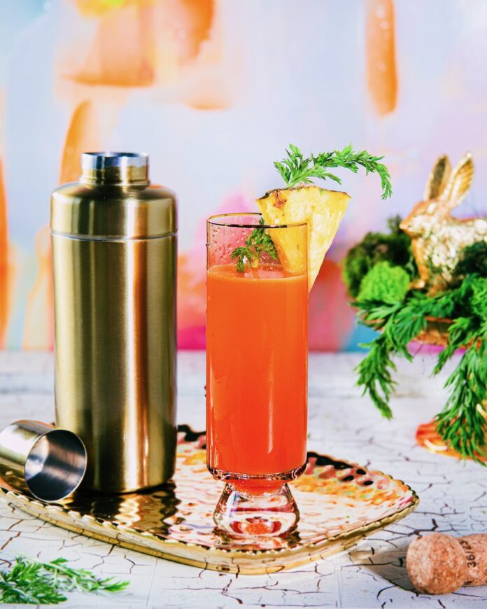 A tall glass holds a Hoppin’ Down the Bunny Trail with a Buzzed Bunny Mimosa in an orange color with a pineapple slice garnish as the whole glass sits beside a gold cocktail shaker.