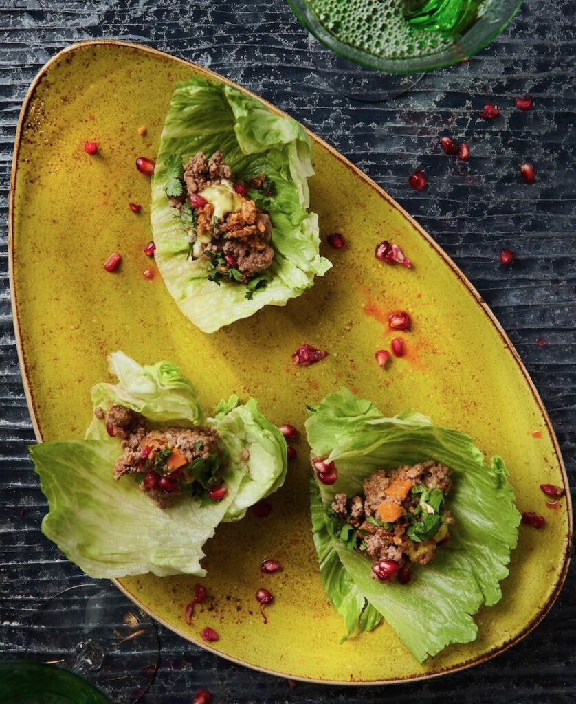 Three iceberg lettuce cups filled with braised lamb, pomegranate seeds, and curry aioli on a yellow plate against a black background.