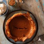 A pumpkin pie with a phyllo crust pits in a pan on a picnic table with small bowls of spices and spoons sitting around it.