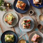 Various light blue and dark blue plates hold different types of focaccia sandwiches like cucumber, cream cheese and salmon, prosciutto, all on a blue and white picnic table.