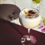 A round glass with a Jennifer Coolidge milk/milf punch cocktail inside a round glass topped with a nutmeg powder and star anise against a flower background.