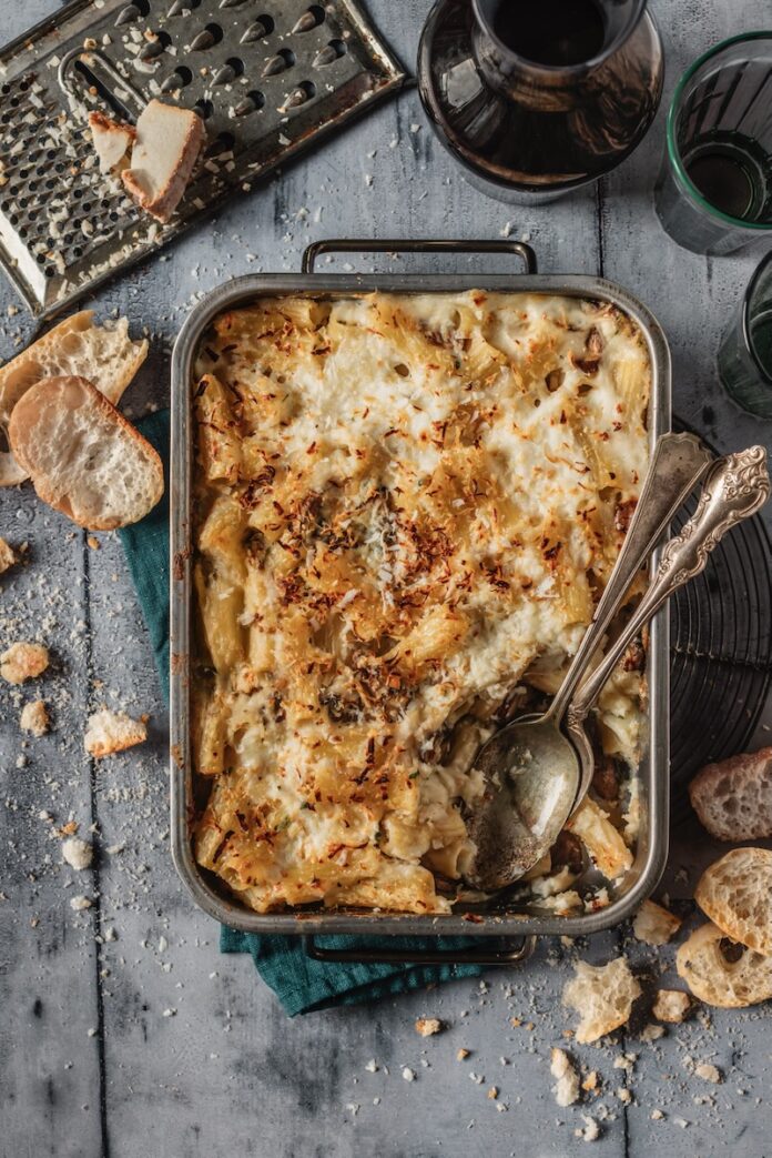 A baking dish with crusty bread sitting around it on a grey table holds a Pasta au Gratin with a spoon scooping out the bottom right corner.