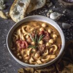 A grey bowl of Pasta e Ceci sits on a black table with pieces of bread and pasta scattered around the bowl.