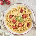 A round white bowl with thick spaghetti inside it featuring halved tomatoes, basil, a Trapanese Pesto and a fork as it sits on a marble countertop.