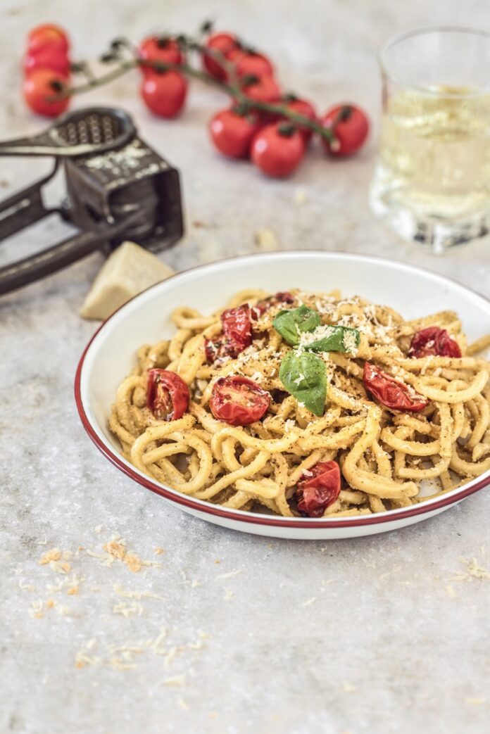A round white bowl holds spaghetti pasta with a Trapanese Pesto on top and halved tomatoes throughout. Basil garnish sits on top.