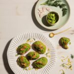A white plate holds a pea puree bruschetta with smoked salmon on top as a green plate off to the side holds a small bowl of the pea purree.