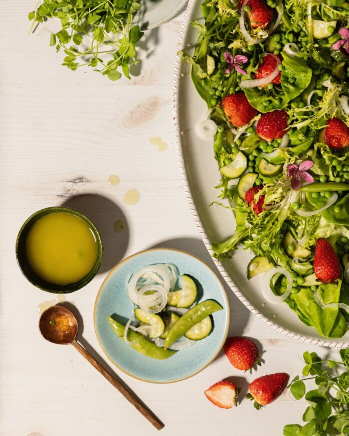 A giant platter of pea salad with strawberries, pickled veggies, and peas sits on a table beside a small bowl of dressing and a blue plate with vegetables on it.