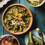 A Persian Noodle Soup sits in a brown bowl on a blue picnic table with three smaller bowls and plates of Persian food sitting around the soup.