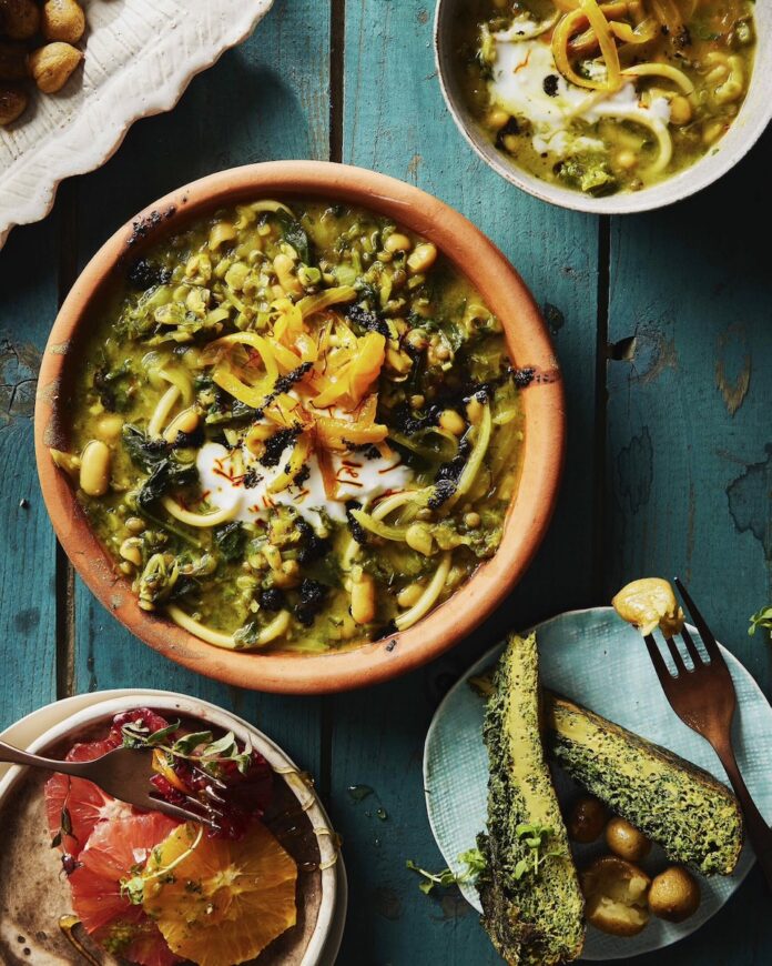 A Persian Noodle Soup sits in a brown bowl on a blue picnic table with three smaller bowls and plates of Persian food sitting around the soup.